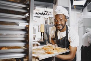 A smiling baker in a white hat and a dark apron holds out a tray of fresh bread beside a rack of baking trays.