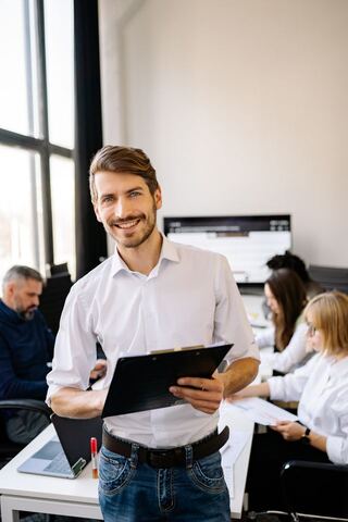 A smiling man in a white shirt holds a clipboard in a bright office, with colleagues working at their desks behind him.
