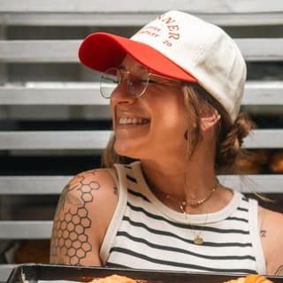A laughing baker in a cap and glasses holds a tray of fresh croissants in front of a bakery rack.