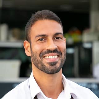 A smiling man in a white shirt and a brown apron stands in front of a café.