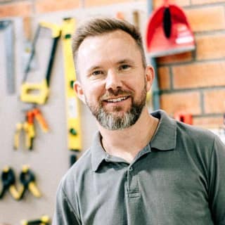 A smiling carpenter leans on his workbench, with tools hanging on the brick wall behind him.