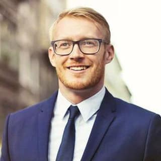 A smiling man in a navy suit and glasses stands on a city street.